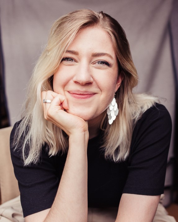 Alina hat Schlüsselbein langes blondes Haar und stützt sich auf ihrer Hand ab, während sie in die Kamera lächelt. Sie trägt silbernen Schmuck und ein schwarzes T-shirt und hinter ihr erkennt man verschwommen einen Fotostudio Hintergrund.