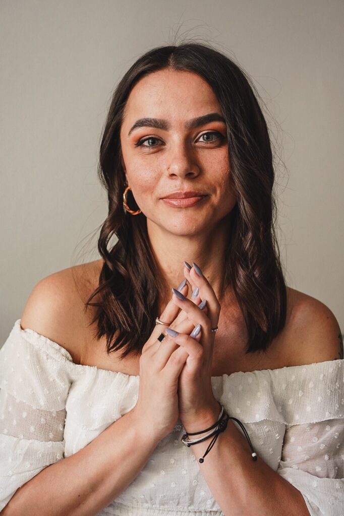 Sabrina trägt ihr Schlüsselbein langes dunkles Haar in dem Bild offen und goldenen Schmuck. Sie lächelt leicht in die Kamera währen, die ihre Hände vor der Brust ineinanderlegt. Ihre Finger sind mit Flieder färbendem Nagellack lackiert und sie trägt eine weiße Bluse, die weiß bepunktet ist.