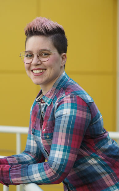 Jennifer, mit kurzem dunklem Haar, welches an der Spitze pink gefärbt ist und Brille, stützt sich mit ihren Ellenbogen auf deinem Geländer ab und lächelt in die Kamera. Der Hintergrund ist verschwommen und sie trägt ein blau rot kariertes Hemd.