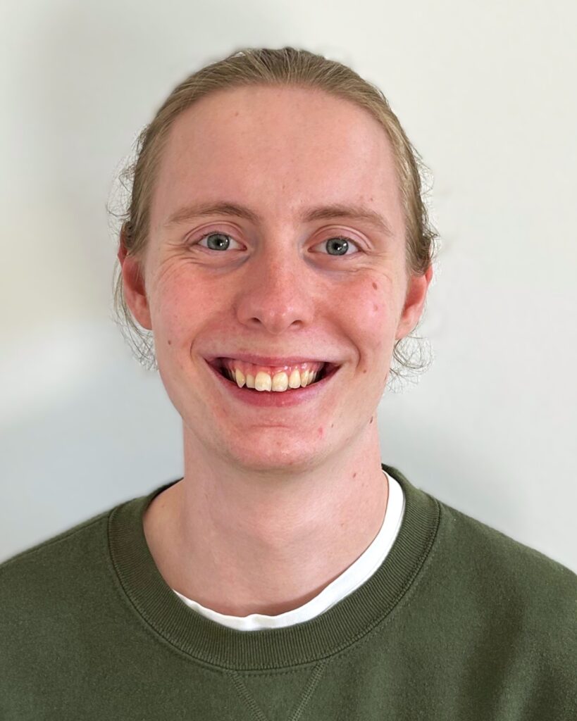 Lorenz trägt seine langen Haare zu einem Dutt und lächelt breit in die Kamera. Er trägt einen Grünen Pullover, unter dem man ein weißes T-Shirt erkennen kann.