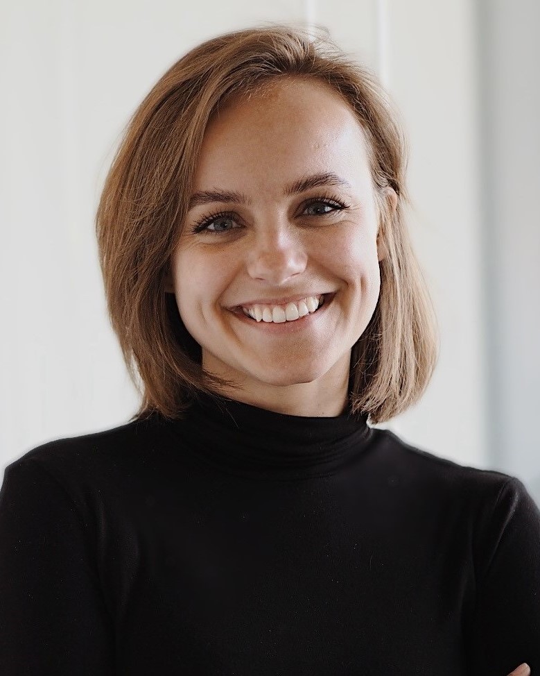 Sophie, trägt einen schwarzen Rollkragen Pullover, hat schulterlanges braunes Haar und lächelt in die Kamera.