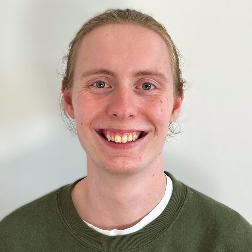 Lorenz trägt seine langen Haare zu einem Dutt und lächelt breit in die Kamera. Er trägt einen Grünen Pullover, unter dem man ein weißes T-Shirt erkennen kann.