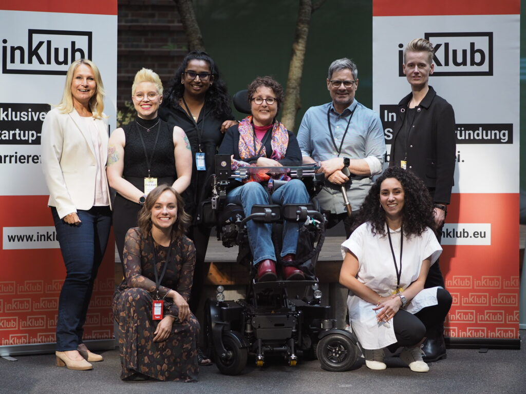 8 Personen stehen nebeneinander und positionieren sich für ein Gruppenfoto. Die Personen sind unterschiedlich, da Frauen und einen Mann, Menschen mit sichtbaren Behinderungen und welche ohne und Personen mit unterschiedlichen Hautfarben im Bild zu sehen sind. Im Hintergrund sieht man an den Ecken jeweils einen inKlub Banner und in der Mitte eine Treppe, die nach oben führt. Alle Personen schauen in die Kamera. Unter den Personen sind die Teilnehmer:innen des Starter Programm, die Jury und Sophie, die Gründerin von inKlub