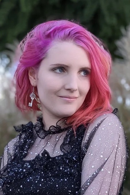Anna posiert mit ihren Pinken Haaren und auffälligen Ohrringen so für die Kamera, dass sie rechts an uns vorbeischaut. Sie lächelt leicht und trägt eine auffällige leicht durchsichtige schwarze Bluse, die mit silberner Farbe bepunktet ist und darunter eine Art schwarzes Top. Im Hintergrund erkennt man unscharf, dass sie sich draußen befindet.