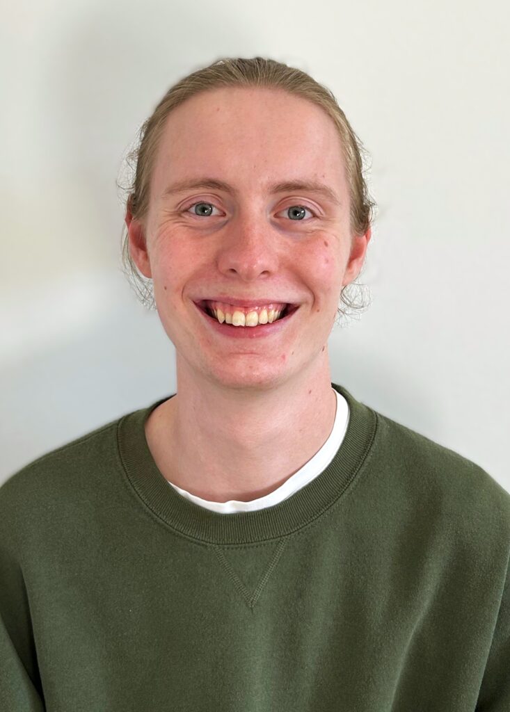 Lorenz trägt seine langen Haare zu einem Dutt und lächelt breit in die Kamera. Er trägt einen Grünen Pullover, unter dem man ein weißes T-Shirt erkennen kann.