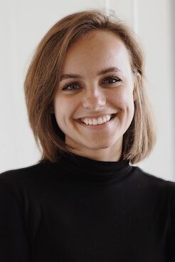 Sophie, trägt einen schwarzen Rollkragen Pullover, hat schulterlanges braunes Haar und lächelt in die Kamera.