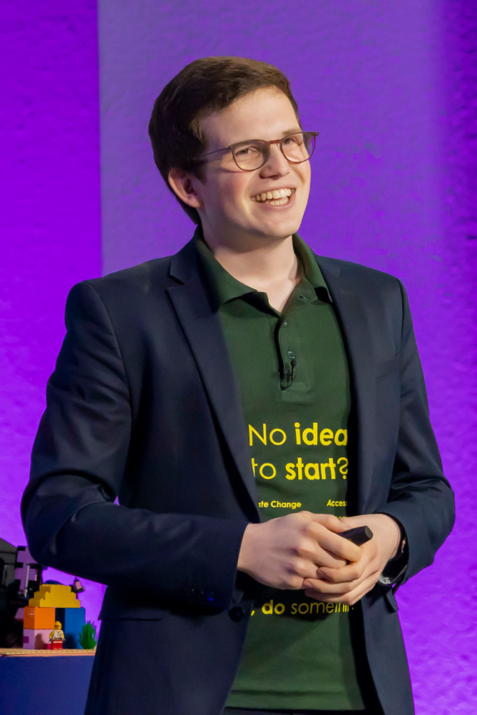 Gernot Jonathan Sümmermann, ein junger Mann in einem dunkelblauen Blazer und einem grünen Polo-Shirt steht lächelnd auf einer Bühne. Das Shirt trägt den Text „No idea to start?“ in gelber Schrift. Der Mann hat braunes, kurzes Haar und trägt eine Brille. In seinen Händen hält er einen Präsentations-Clicker. Der Hintergrund ist in einem intensiven Lila beleuchtet, und im Vordergrund ist ein LEGO-Modell zu erkennen.