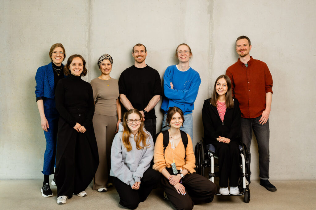 9 Personen, mit teilweise sichtbaren Behinderungen, stehen zum Gruppenbild und schauen freundlich in die Kamera