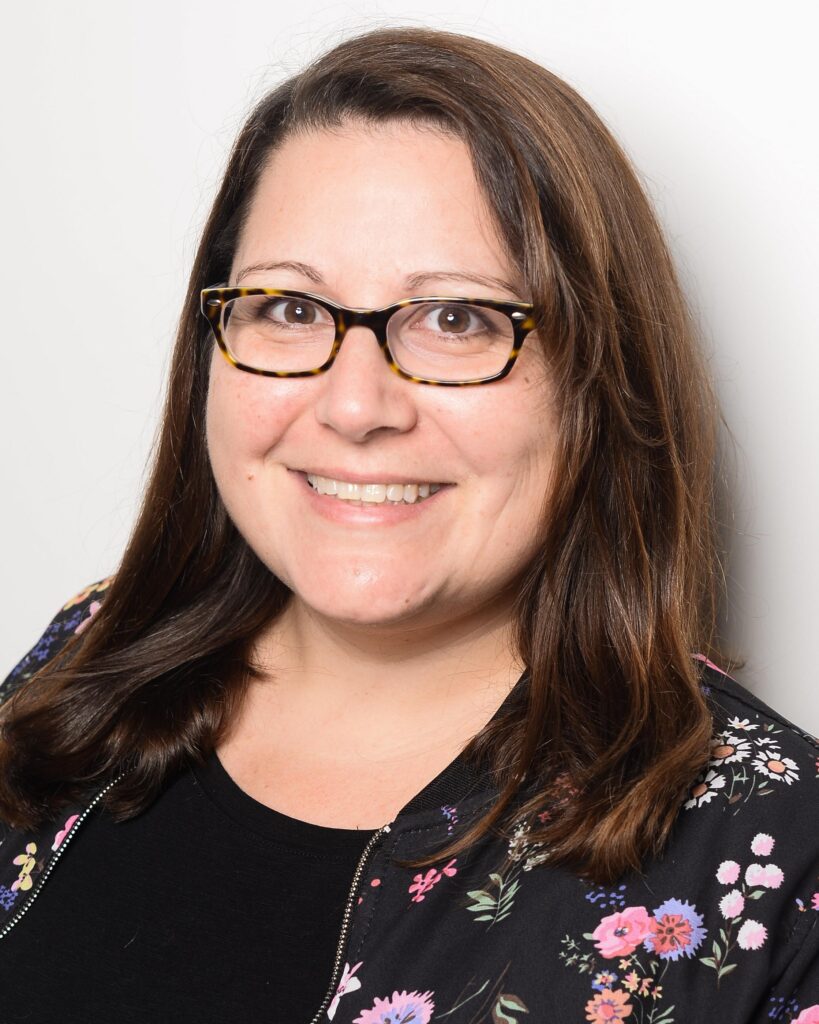 Yasmin trägt ihr Schlüsselbein langen dunklen Haare offen und lächelt breit in die Kamera. Sie hat eine Brille auf und schwarze Jacke auf der Blumen abgebildet sind und darunter ein schwarzes T-Shirt.