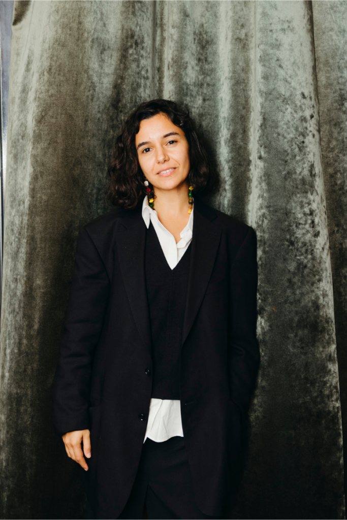 Sema Gedik, eine Person mit schulterlangem dunklem Haar steht vor einem samtartigen, grün-grauen Vorhang. Sie trägt ein schwarzes Outfit bestehend aus einem Blazer, einer Weste und einer weißen Bluse. Sie lächelt leicht und trägt farbige Ohrringe