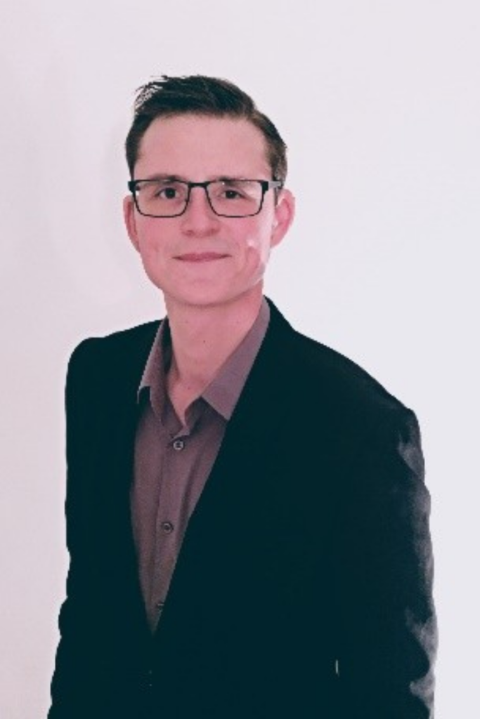 Alexander lächelt in die Kamera. Er trägt eine Brille, kurzes dunkles Haar und ein schwarzes Jacket über einem braunen Hemd.