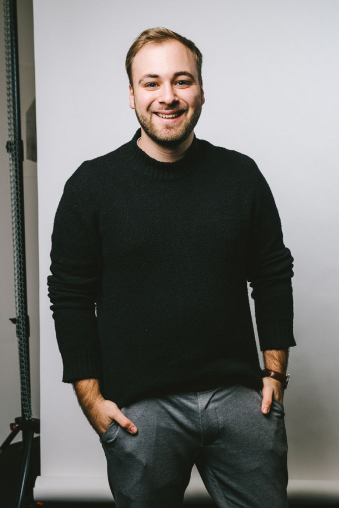 Lukas lächelt in die Kamera. Er trägt einen Schwarzen Pullover und eine dunkle Hose, einen kurzen Bart und kurze Haare.