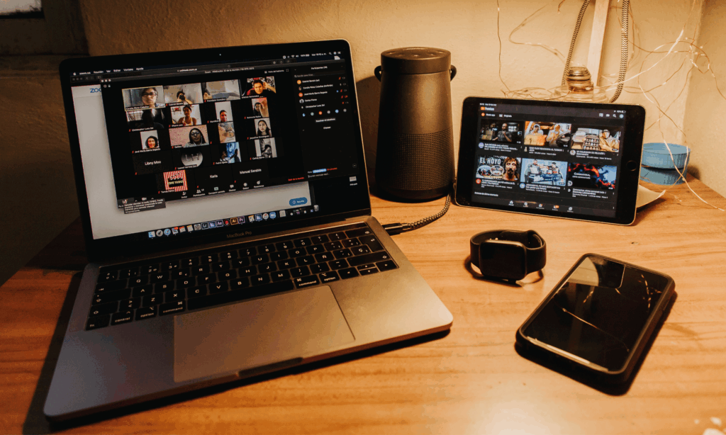 Auf einem Laptop sieht man ein offenes Teams Meeting mit vielen unterschiedlichen Personen. Neben dem Laptop steht ein Tablet ein Handy und eine Smartwatch.